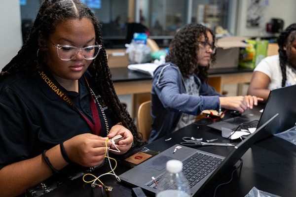 Two students engaged in a hands-on technical activity at a summer camp, working with wires and a laptop at a lab table.