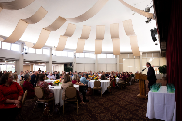 A speaker at a podium addressing an audience seated at round tables during an event in a conference room.