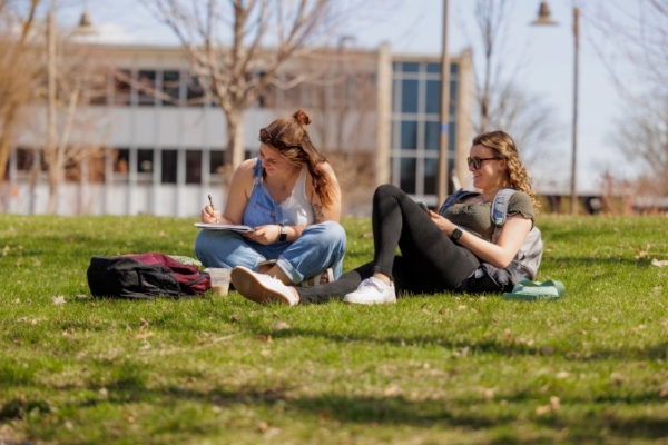 Two students sitting on the grass outdoors on a sunny day; one is writing in a notebook, and the other is looking at a phone with a blurred building at backside.