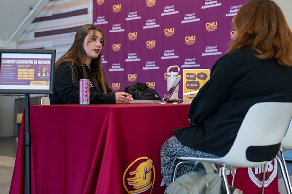 Two women seated at a booth for Career Coaching in CMU at an event, with a branded purple backdrop featuring the College of Business Administration logo and a red tablecloth.