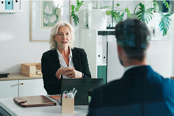 A woman with short white hair wearing a black sweater and white blouse talks to a man in a suit coat.