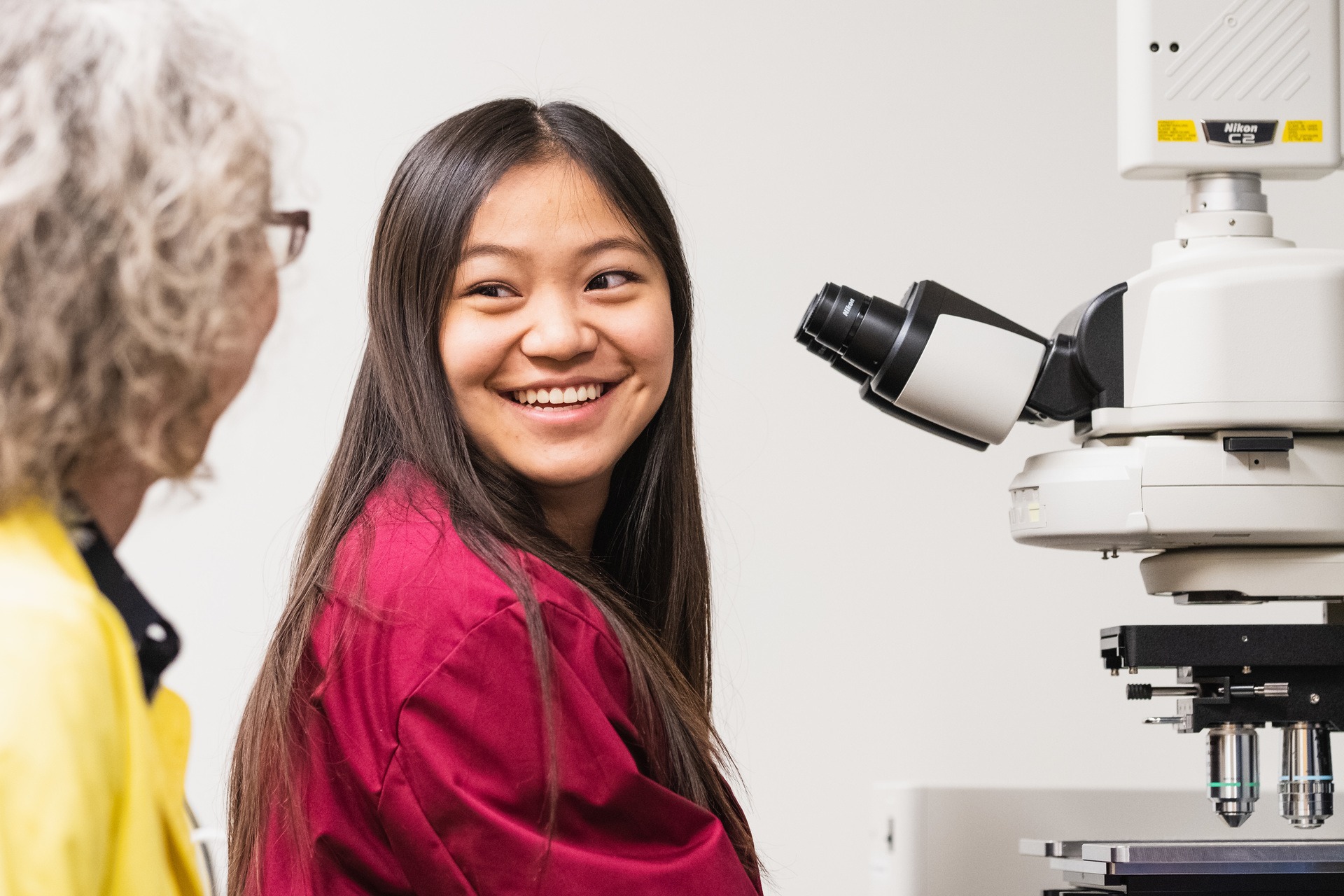 Student sitting in front of a microscope looking at a woman with curly grey hair.