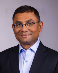 Trustee Kondur A man in a dark suit smiling in front of a light grey background.