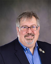 Professional headshot of Trustee Todd J. Regis in a blue suit in front of a grey background.