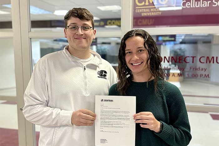 Jared (left) is presented with his scholarship award letter by Ashley (right). The two are standing in front of a glass wall with the CONNECT CMU logo on it.