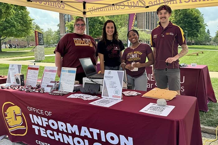 Four OIT staff members stand behind a table under a tailgate tent. Each is wearing CMU shirts.
