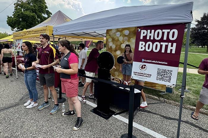 Three OIT staff members encourage passersby to stop into the OIT photo booth.  Staff are standing in front of the tent with booth in the background.