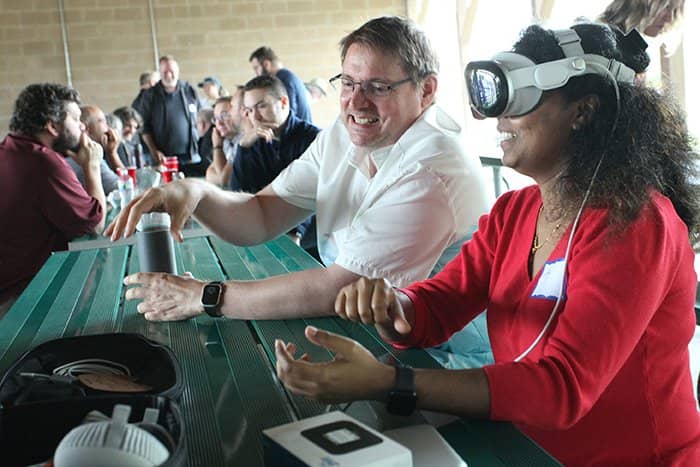 An OIT staff member in a white shirt demonstrates the Apple Vision Pro device for a colleague who is earing a read shirt.