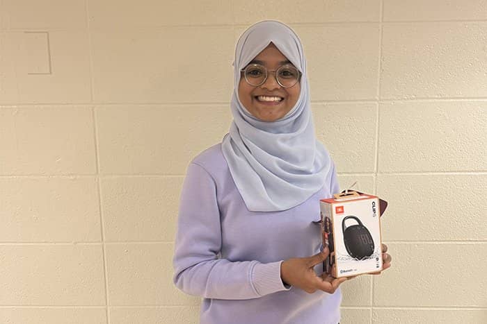 Ayehsa poses with a speaker while wearing lavender shirt and head wrap and standing in front of a cream colored wall.