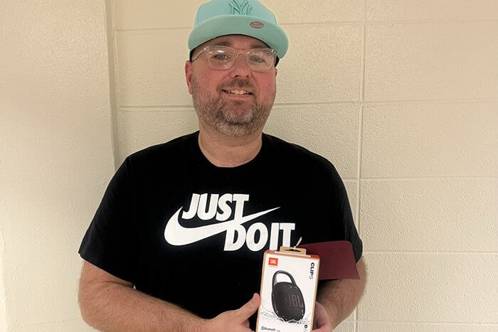 Carl poses with a speaker while wearing a black t-shirt and teal NY Yankees hat and standing in front of a cream colored wall.