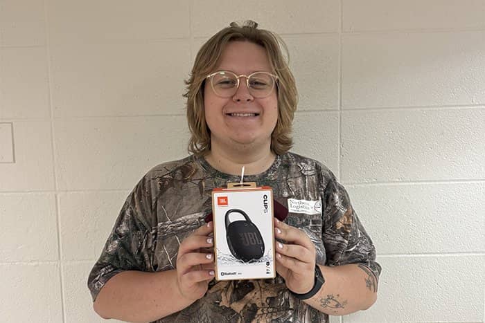 Tyler poses with a speaker while wearing a camouflage shirt and standing in front of a cream colored wall.