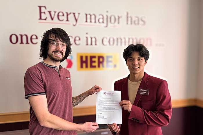 Miles presents a scholarship award letter to Hasanain while standing in front of a sign that reads Every major has one place in common... HERE.