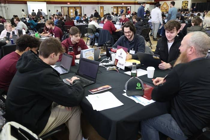 Students sit at a table and work on laptops while their advisor looks on.