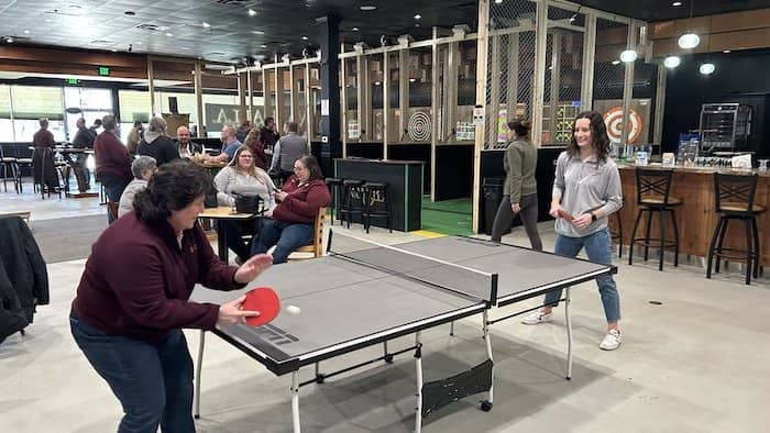 Two staff members play ping pong while others throw axes and visit at tables in the background.