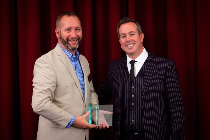 Portrait of Brian Roberts holding his award alongside CMU President Neil MacKinnon. Brian is in a tan jacket and blue shirt and Mackinnon in a dark suit with light pinstripes.
