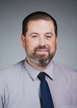 Tim Gramza smiles for a professional headshot and wears a mauve shirt with a dark blue tie. He stands against a gray background.