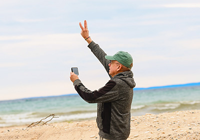 A man at the beach holding up three fingers while holding his phone in his other hand