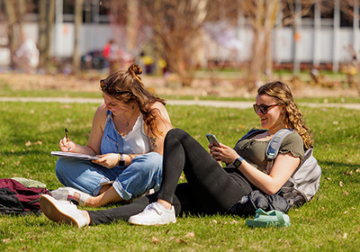 Two students sitting in the grass on campus, one writing in a notebook and the other on her phone