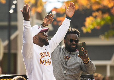 Two men in CMU gear, one with both hands raised and the other with one hand up while holding a phone