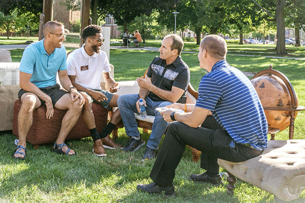 Four men are outside on a sunny day sitting on and among an assortment of furniture that has been set out on the grass for CMU's Get Acquainted Day.