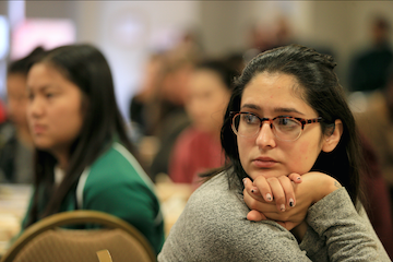 A student sits at a round table listening intently to a keynote speaker in large event room.