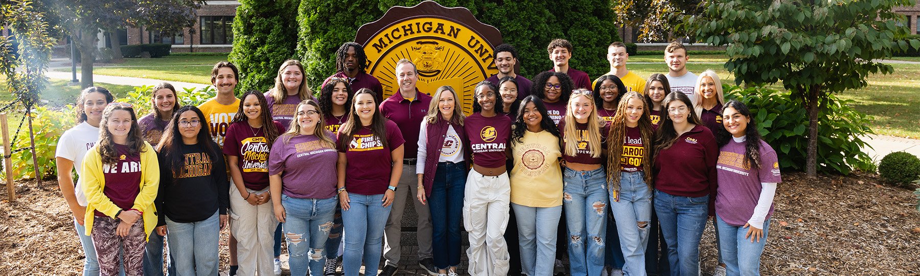 A group of Central Michigan students stand with Dr. MacKinnon and his wife in front of the CMU seal in Warriner Mall.