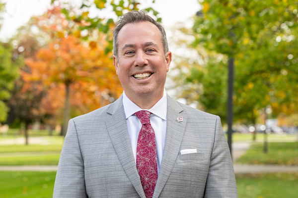 Professional headshot of President Neil MacKinnon wearing a gray suit smiling at the camera outdoors with trees in the background.