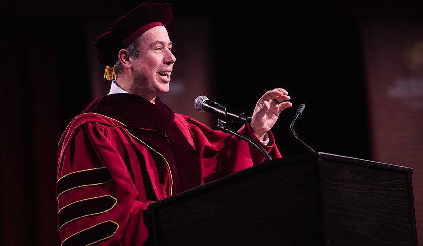 A man in a red academic gown speaks into a microphone.