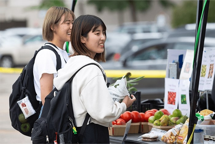 Two students smiling and standing near a produce booth at a farmers market.
