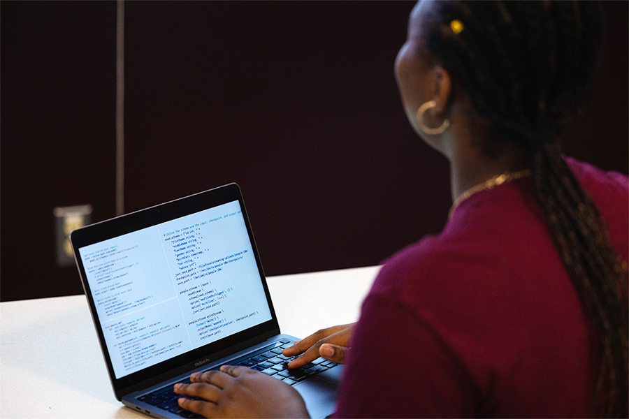 A student in maroon with gold earrings and gold necklace, typing code on a laptop.