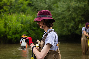 Male student in waders and CMU hat takes water samples from the Chippewa River near Central Michigan University campus.
