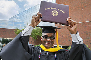 A very happy African-American man in full graduation regalia holds his diploma up for the camera.