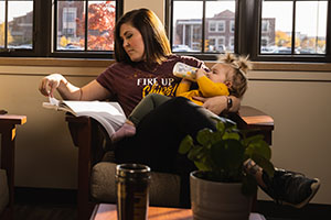 Mother sits in chair reading from a textbook while bottle feeding her baby.