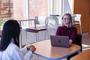 A professor sits at a desk with a computer on the table talking to a student.