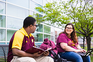 Two students talk on a bench in front of a building