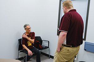 a man holding his shoulder while sitting in a chair with a man giving him an exam standing in front of him