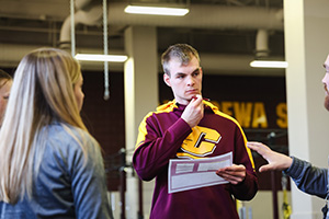 a man holding a piece of paper and wearing a CMU sweatshirt while talking to a woman and man standing in front of him