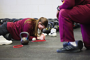 a woman doing a plank on the floor with weights surrounding her and someone kneeling to watch in the foreground