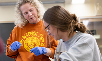 A professor wearing a shirt that reads 'CMU Biological Station' with the CMU seal has blue latex gloves on looking down at a student's work in an anatomy class.