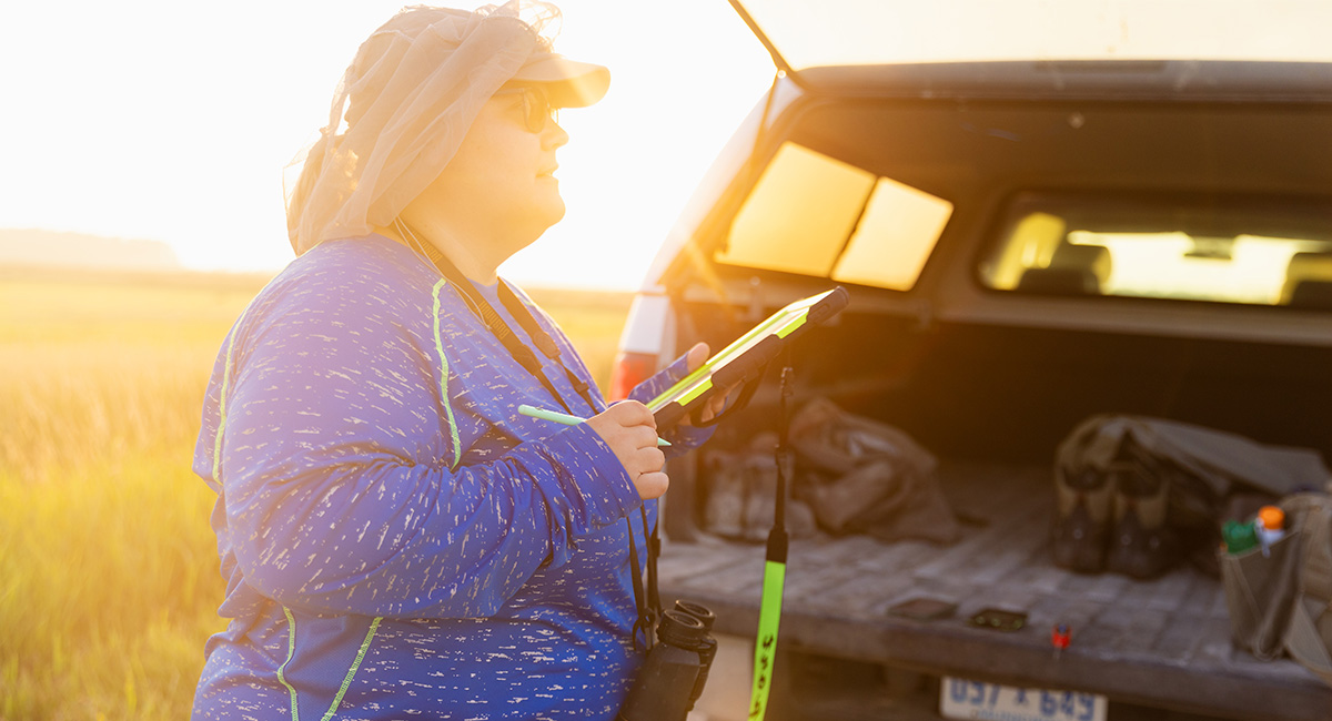 Biotech-1200x650 A student in a blue shirt, sunglasses and a netting hat, holds a clipboard standing behind the bed of a truck.