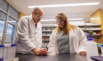 biotech-350x210 A man in a white lab coat and safety glasses looks at a petri dish next to a blonde woman in a white lab coat.