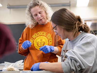 biotech-400x300 A woman in an orange shirt wearing blue latex gloves works with a student wearing glasses and blue gloves.