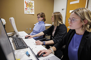 Two students working on computers with a woman sitting in front of a hearing monitor.