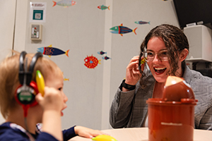 A student wears headphones and works with a young boy who also has headphones on.
