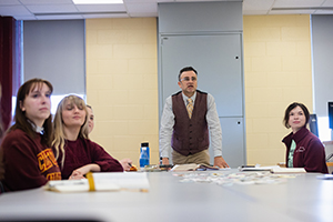 A man standing in front of a group of people sitting at a table while they stare past the camera observing something.