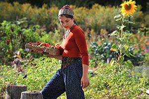A woman wearing a scarf on her head and wearing an orange sweater walks through a garden carrying a basket of produce.