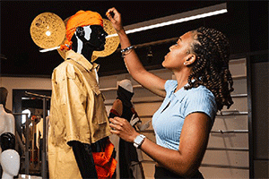 A fashion student dresses a mannequin to wear a tan shirt and orange head piece.