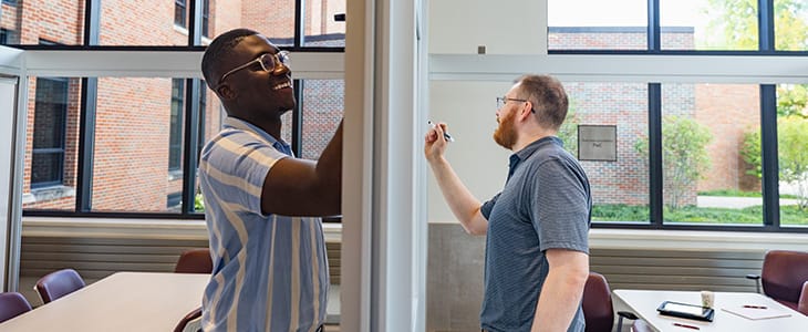 Two men wearing glasses work on opposite sides of a white board.