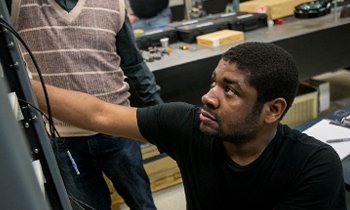 A student working on a science project with another student behind him wearing a sweater vest.