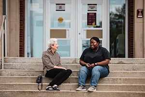 two women sitting on steps in front of CMU building laughing with each other as they have a conversation